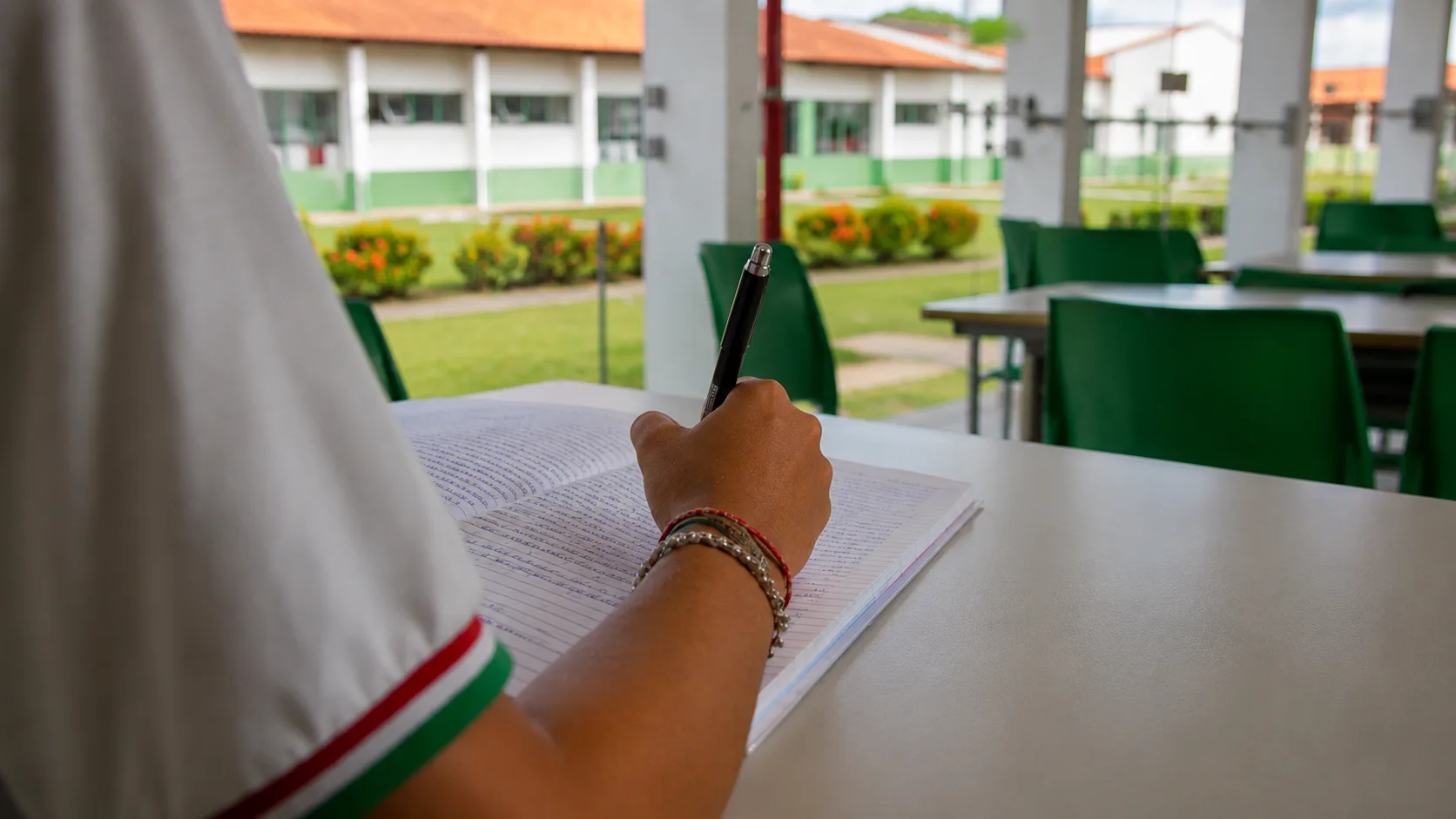 Estudante escreve uma redação em um caderno no IF Baiano, com o campus desfocado ao fundo.
