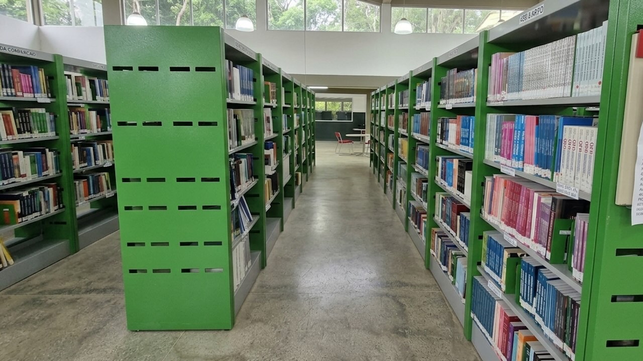 Fotografia de um corredor central de biblioteca com estantes verdes repleta de livros. Ao fundo, uma janela ampla revela árvores e iluminação natural sob um teto elevado com luminárias pendentes.