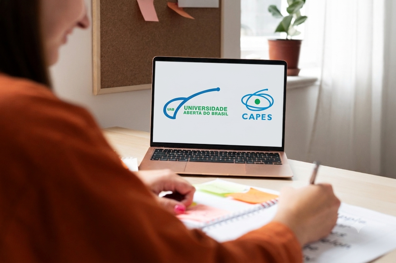 Imagem de uma pessoa escrevendo em uma mesa com um notebook aberto à frente. A tela do notebook exibe os logotipos da UAB Universidade Aberta do Brasil e da CAPES.