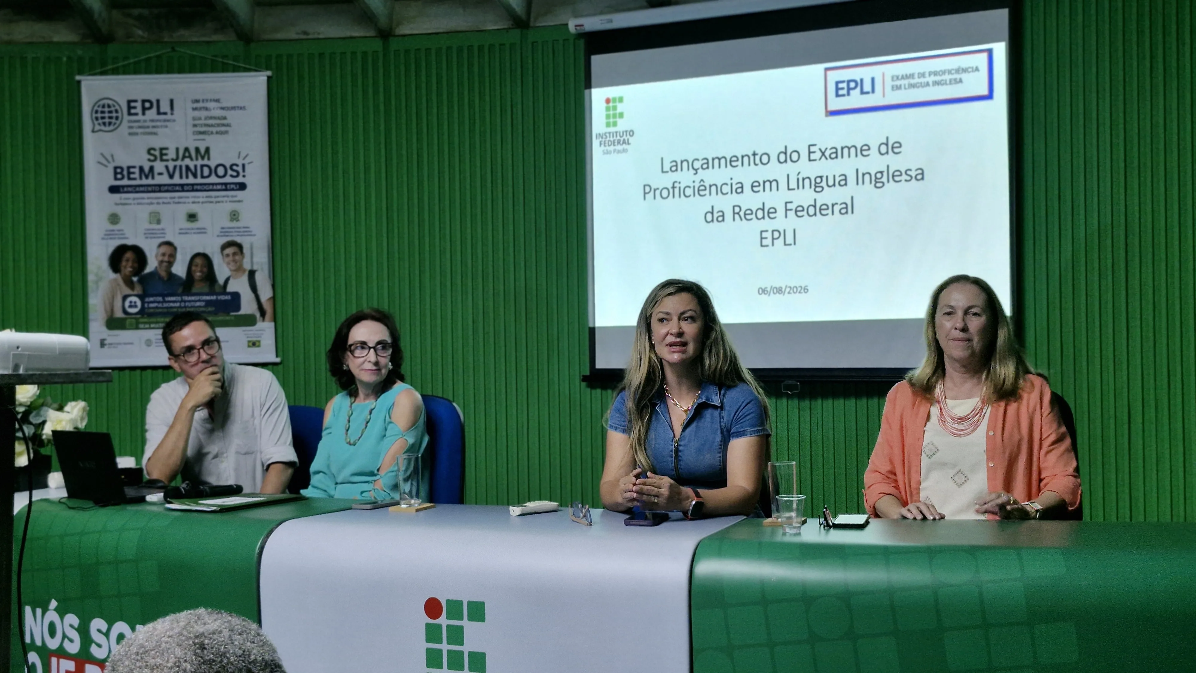 Painel com quatro pessoas sentadas à mesa durante um evento do Instituto Federal Baiano. Ao fundo, um telão apresenta o lançamento do Exame de Proficiência em Língua Inglesa da Rede Federal (EPLI), datado de 6 de agosto de 2026. O público acompanha a apresentação em primeiro plano.